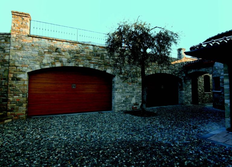 Basculante in legno rosso mogano su foro ad arco in muro di pietra, cortile con ulivo