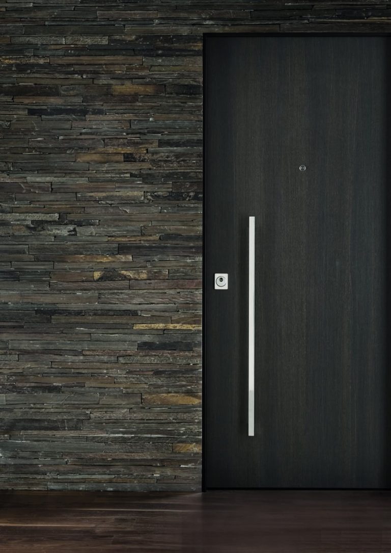 Porta blindata scura con maniglione verticale su parete in pietra a listelli (verticale)